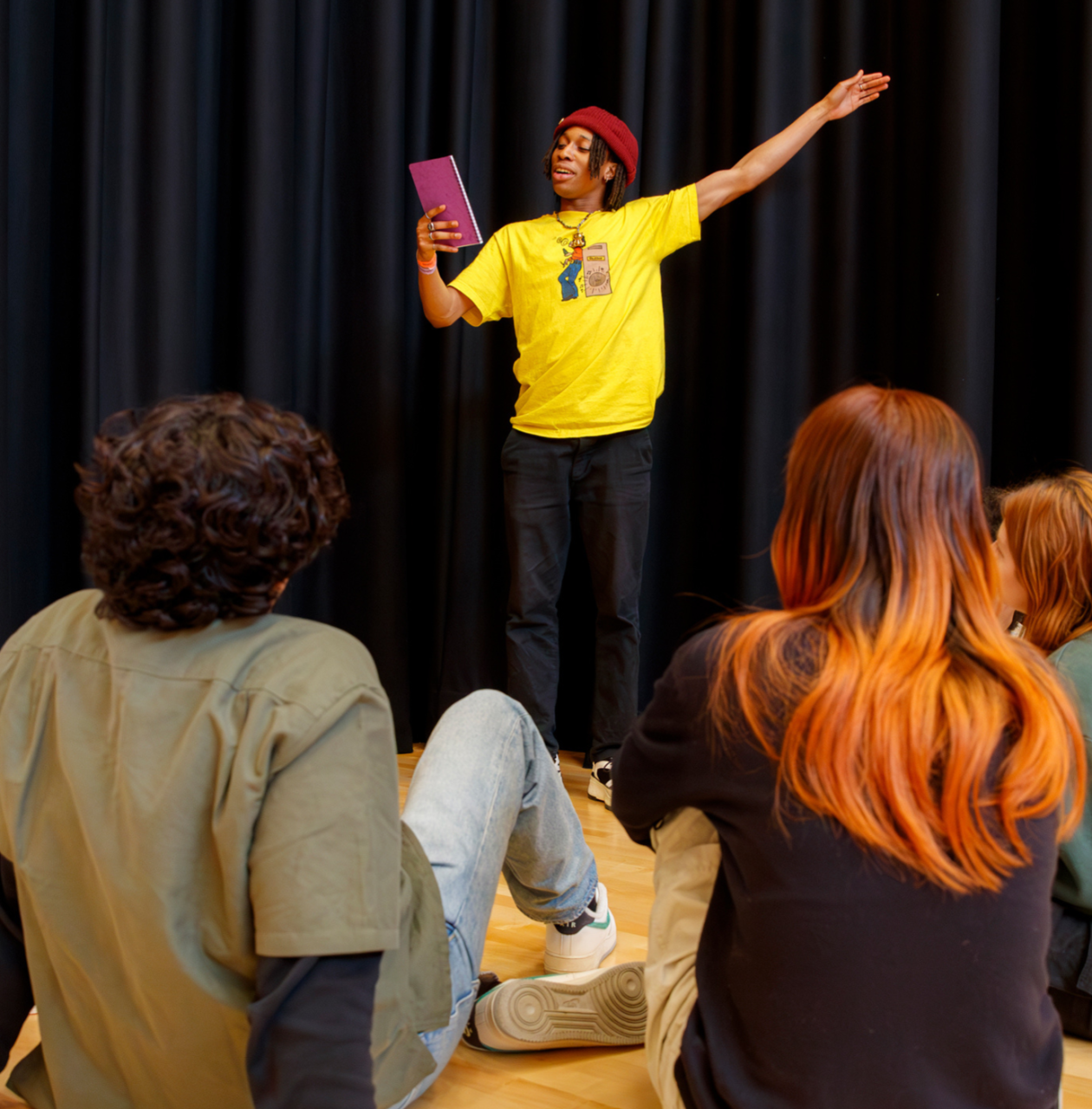 A young person in a yellow shirt performs a monologue to a group of other young people sat on the floor around him