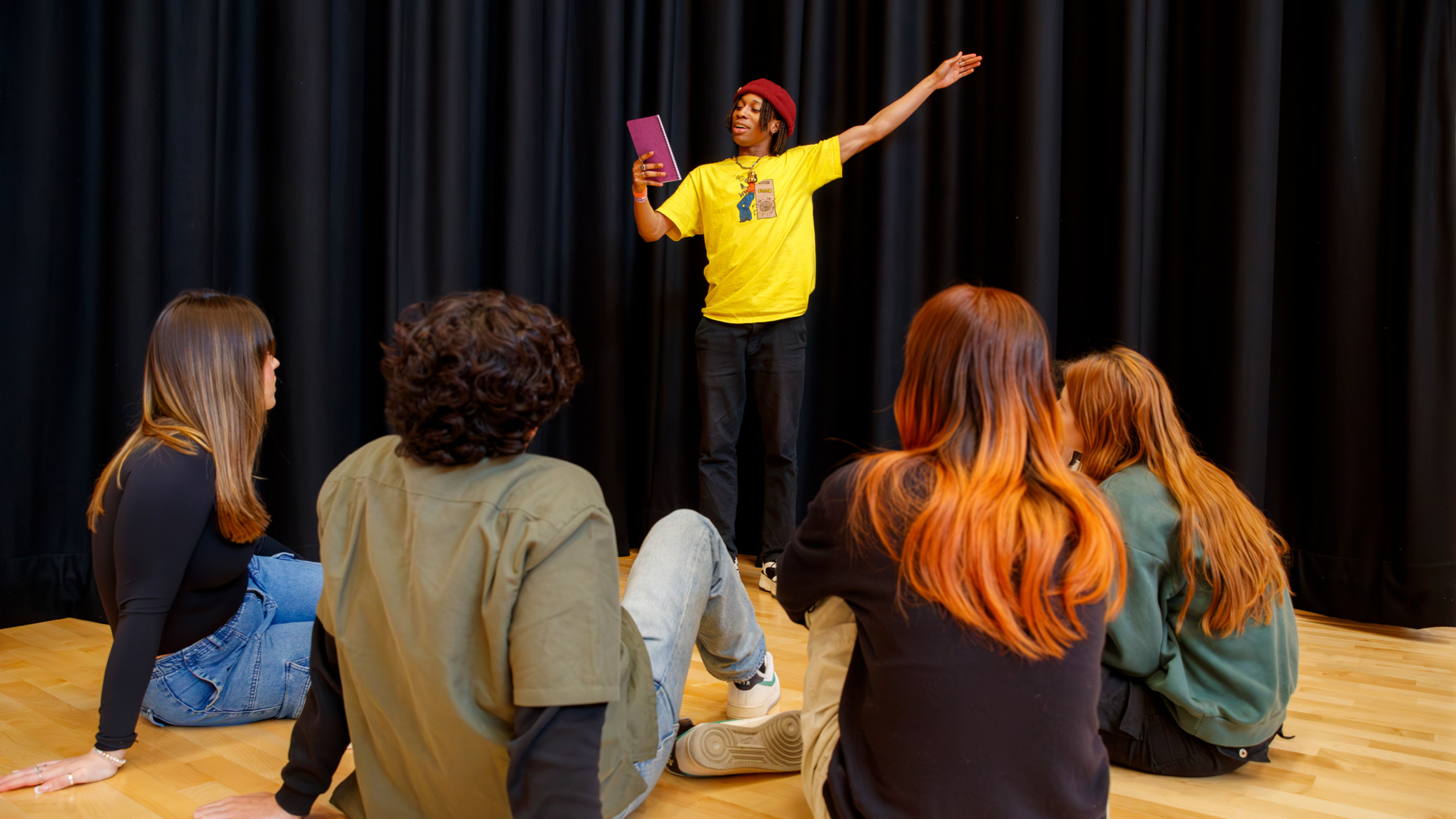 A young person in a yellow shirt performs a monologue to a group of other young people sat on the floor around him