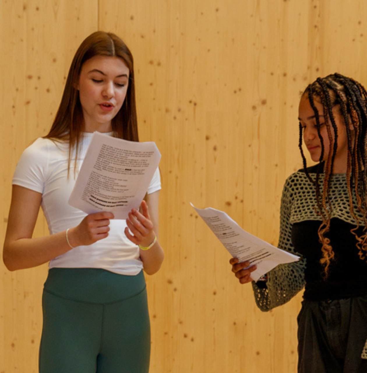 wo young women stand in front of a wooden wall, holding scripts and performing a scene together.