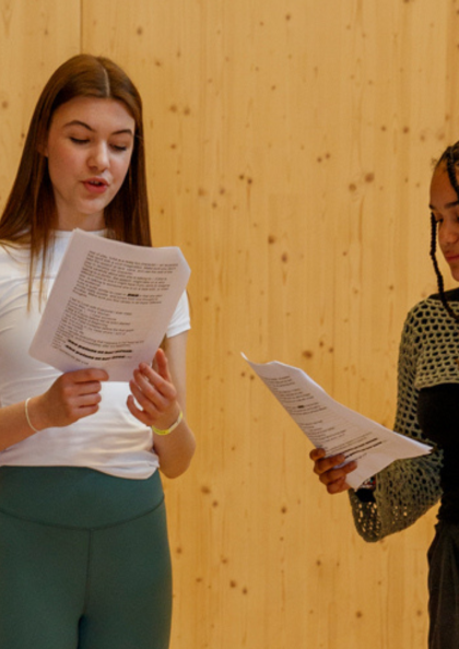 wo young women stand in front of a wooden wall, holding scripts and performing a scene together.