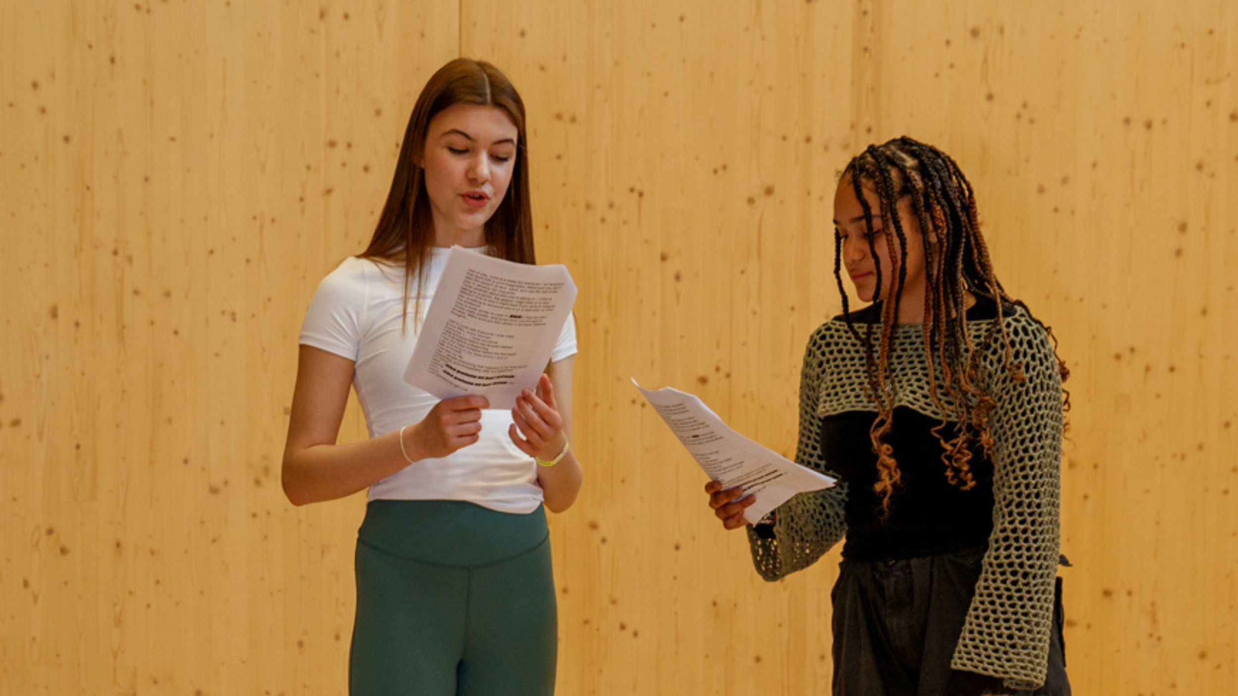 wo young women stand in front of a wooden wall, holding scripts and performing a scene together.