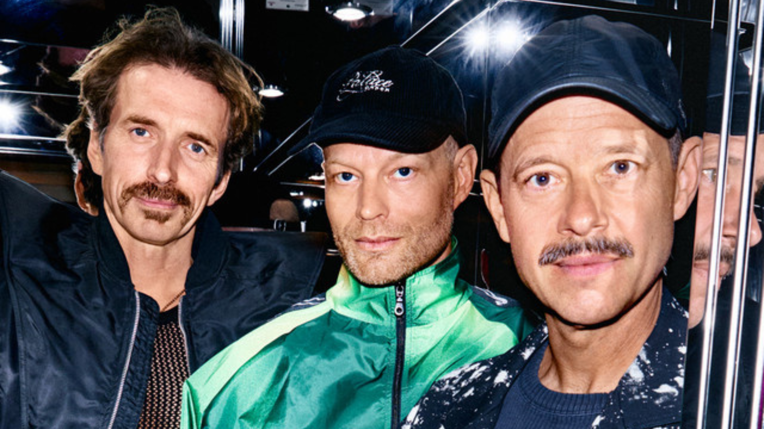 e Danish electronic trio WhoMadeWho pose closely together in a vibrant, high-contrast press photo, with reflective surfaces and intense lighting enhancing their bold, modern aesthetic.
