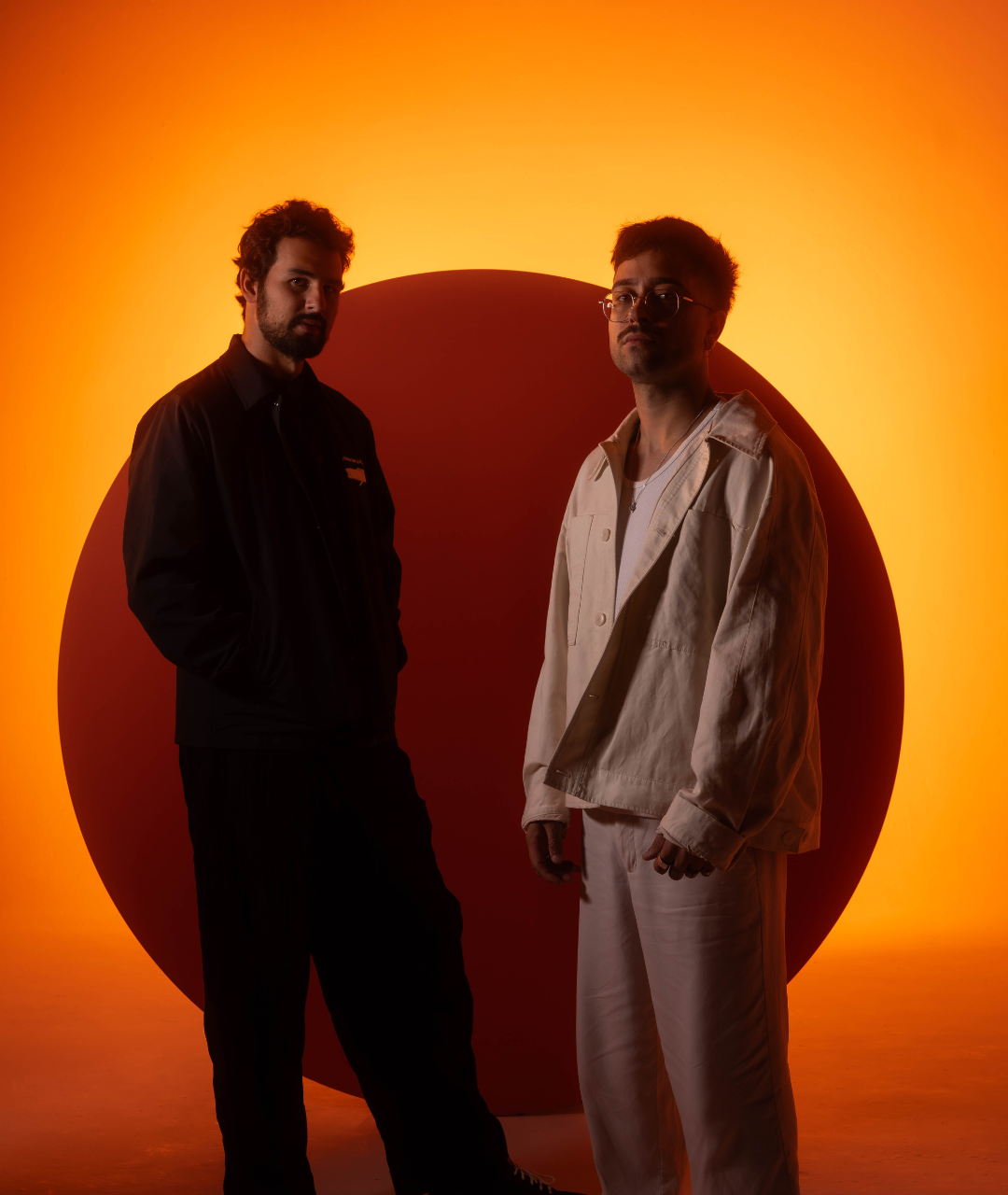 Maz & Antdot standing in front of a glowing orange and red backdrop, wearing dark and light jackets respectively, in a stylized press photo.