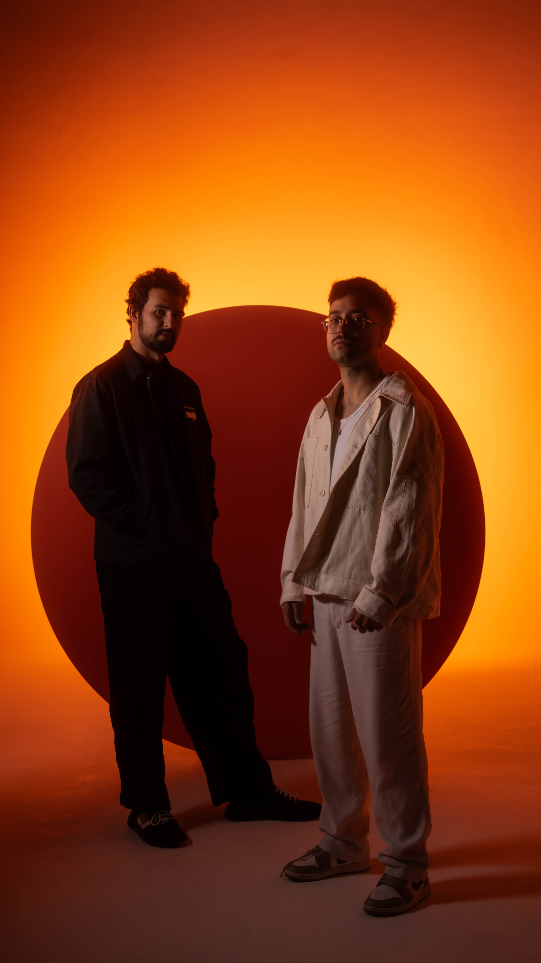 Maz & Antdot standing in front of a glowing orange and red backdrop, wearing dark and light jackets respectively, in a stylized press photo.