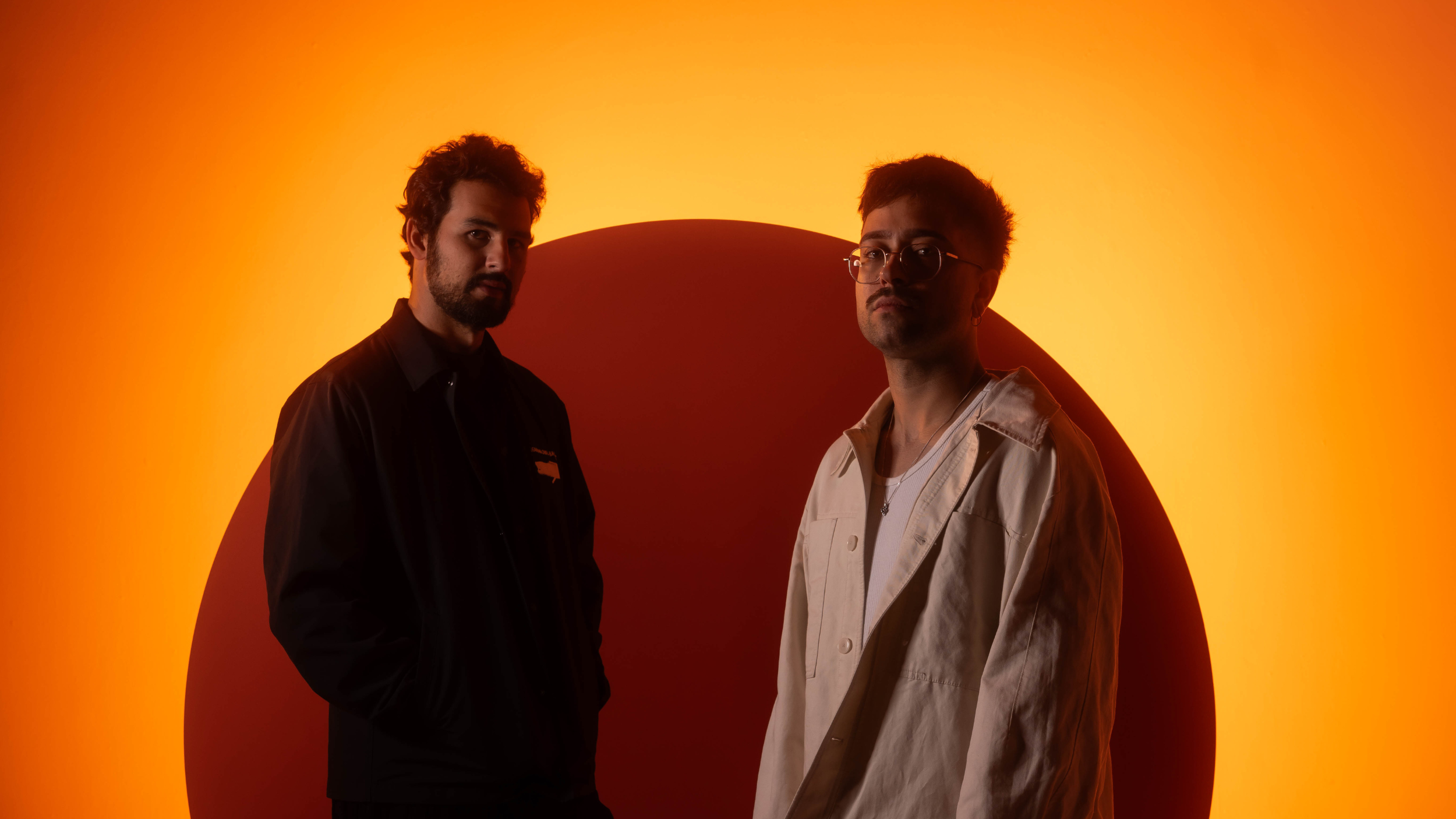 Maz & Antdot standing in front of a glowing orange and red backdrop, wearing dark and light jackets respectively, in a stylized press photo.