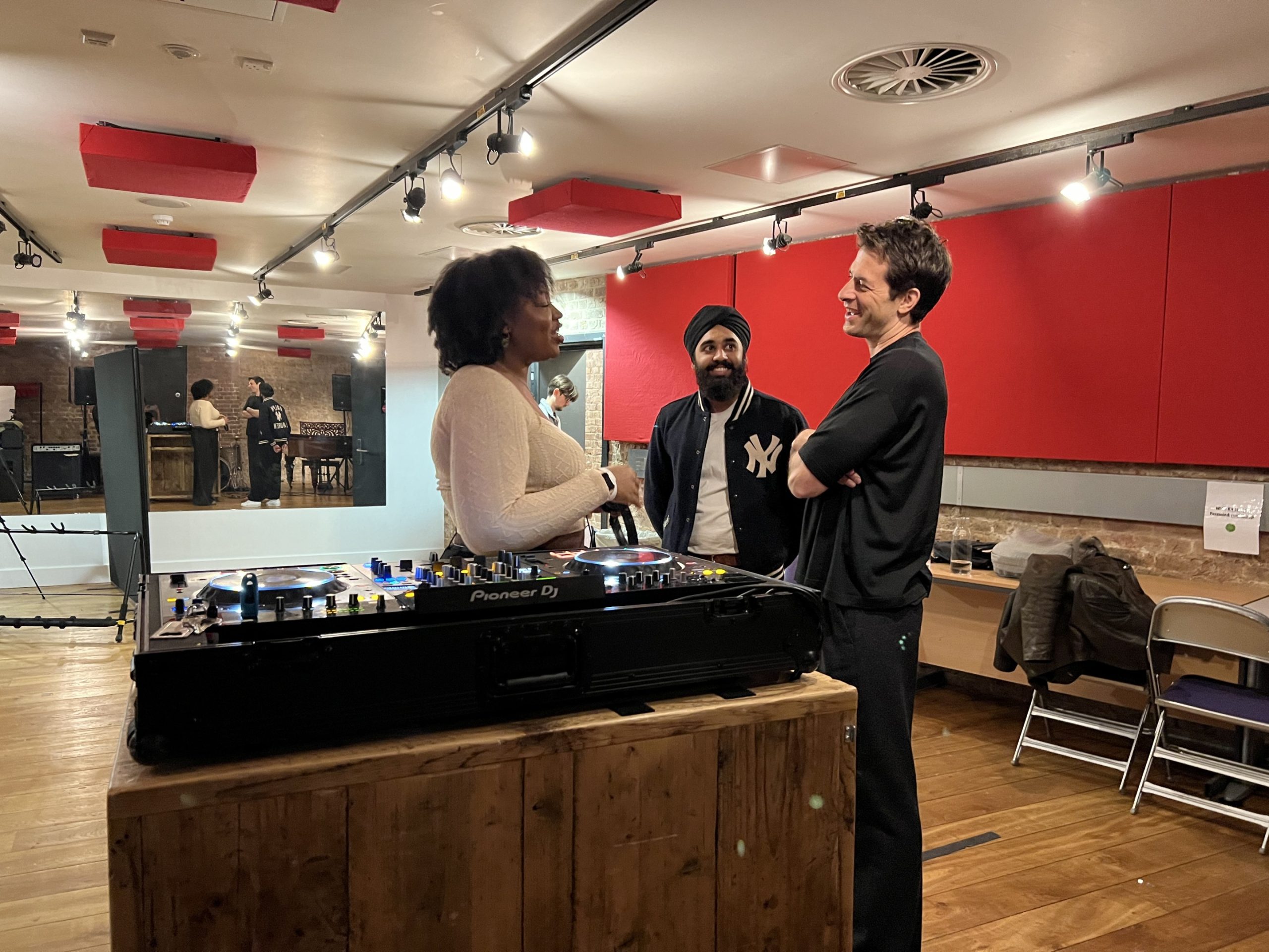 Three people chat in front of a DJ desk in a studio room.