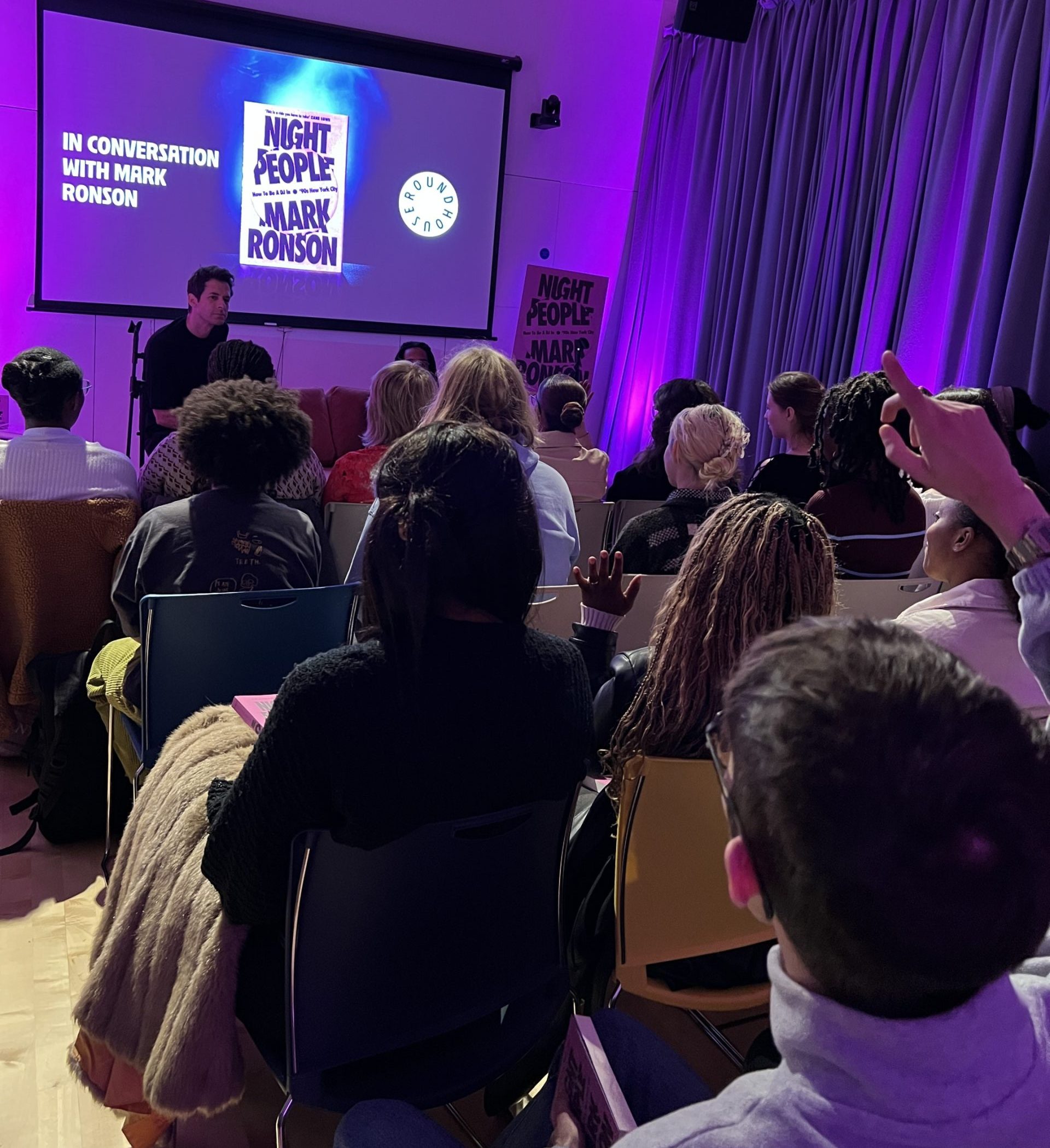 A group of people sit in chairs facing a projector, which reads 'In Conversation with Mark Ronson' and has a picture of his new book Night People