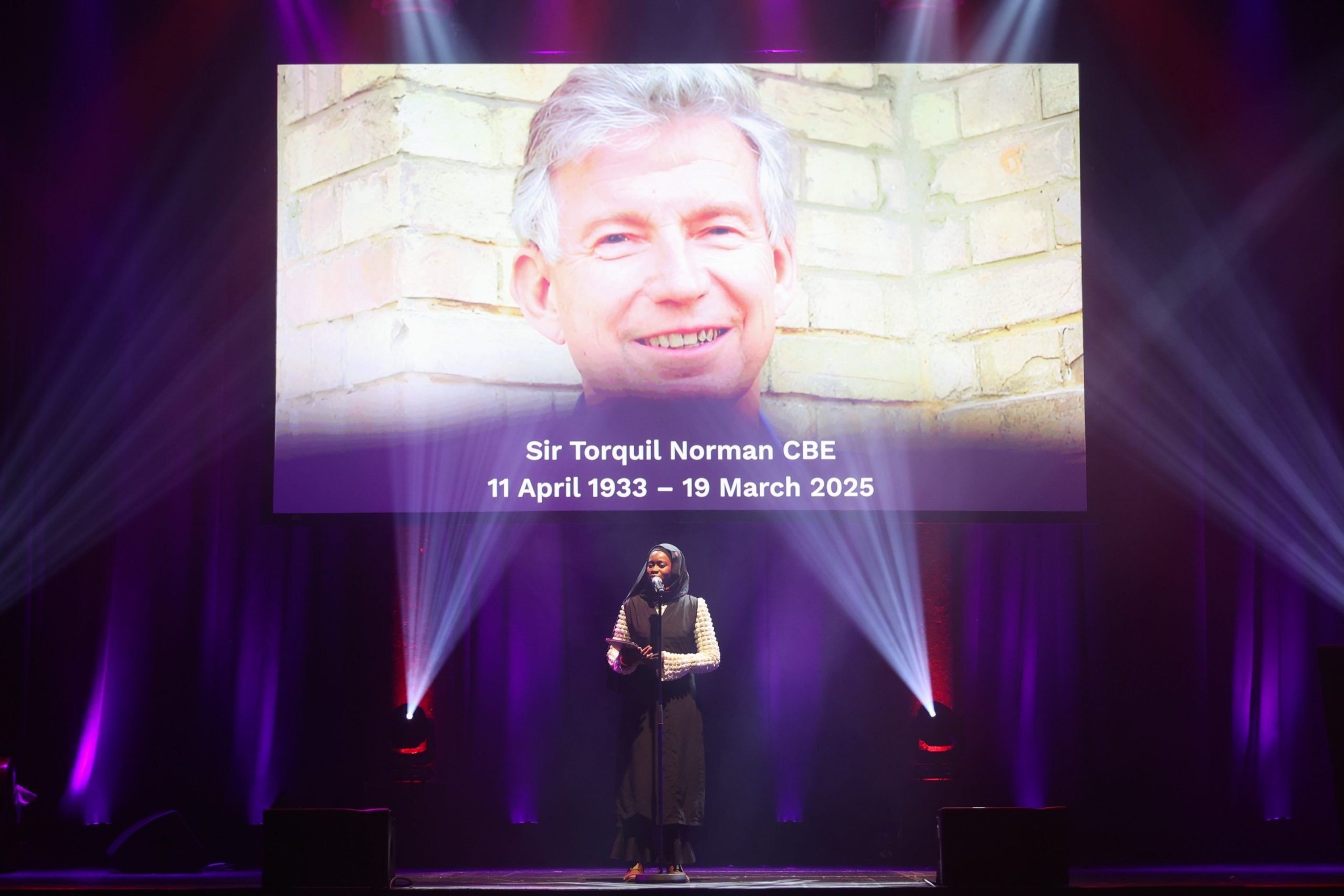 A woman stands in front of a projection of a photo of a man. The photo reads 