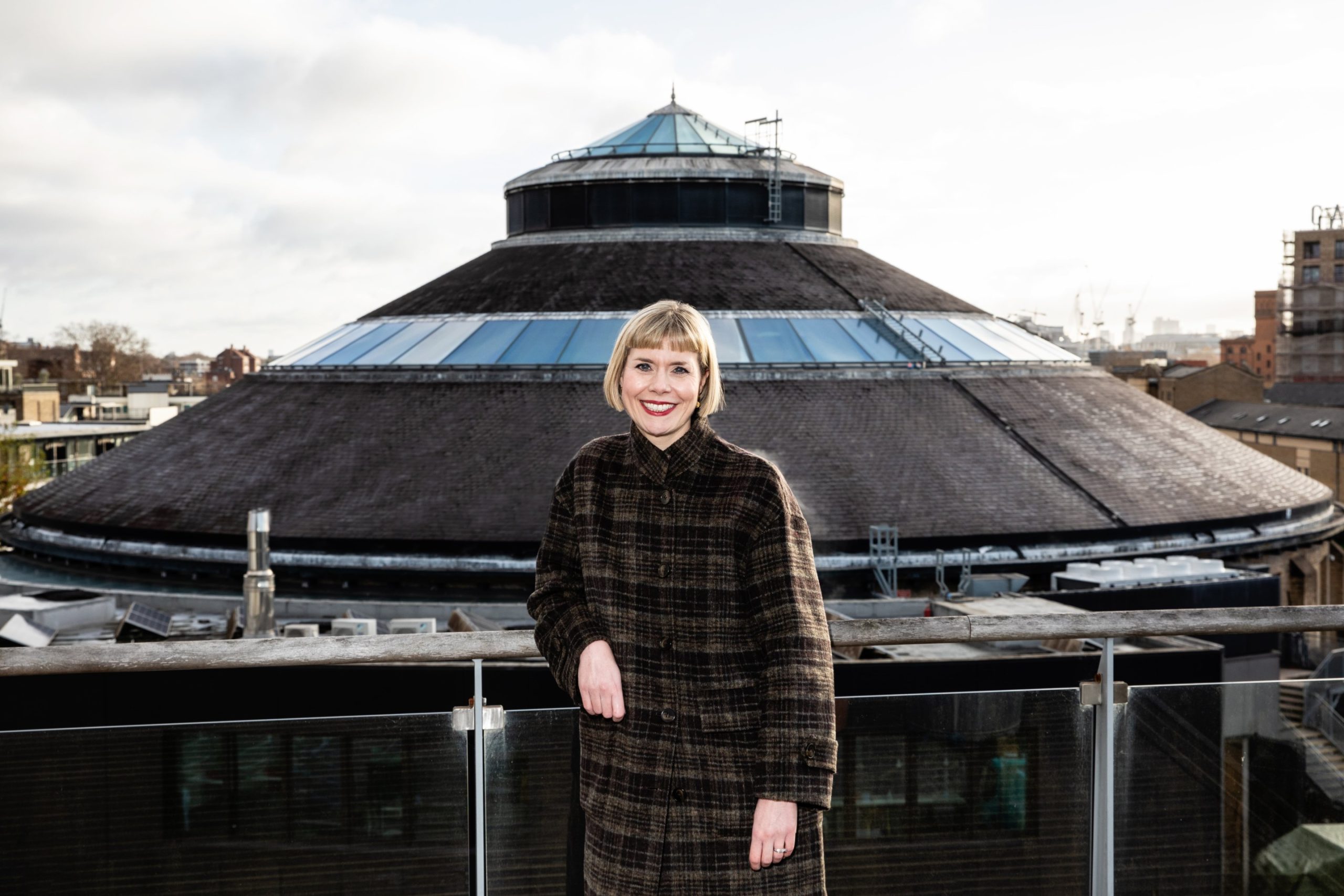 A woman in a brown coat with a blonde bob stands on a balcony in front of the pointed roof of the Roundhouse music venue.
