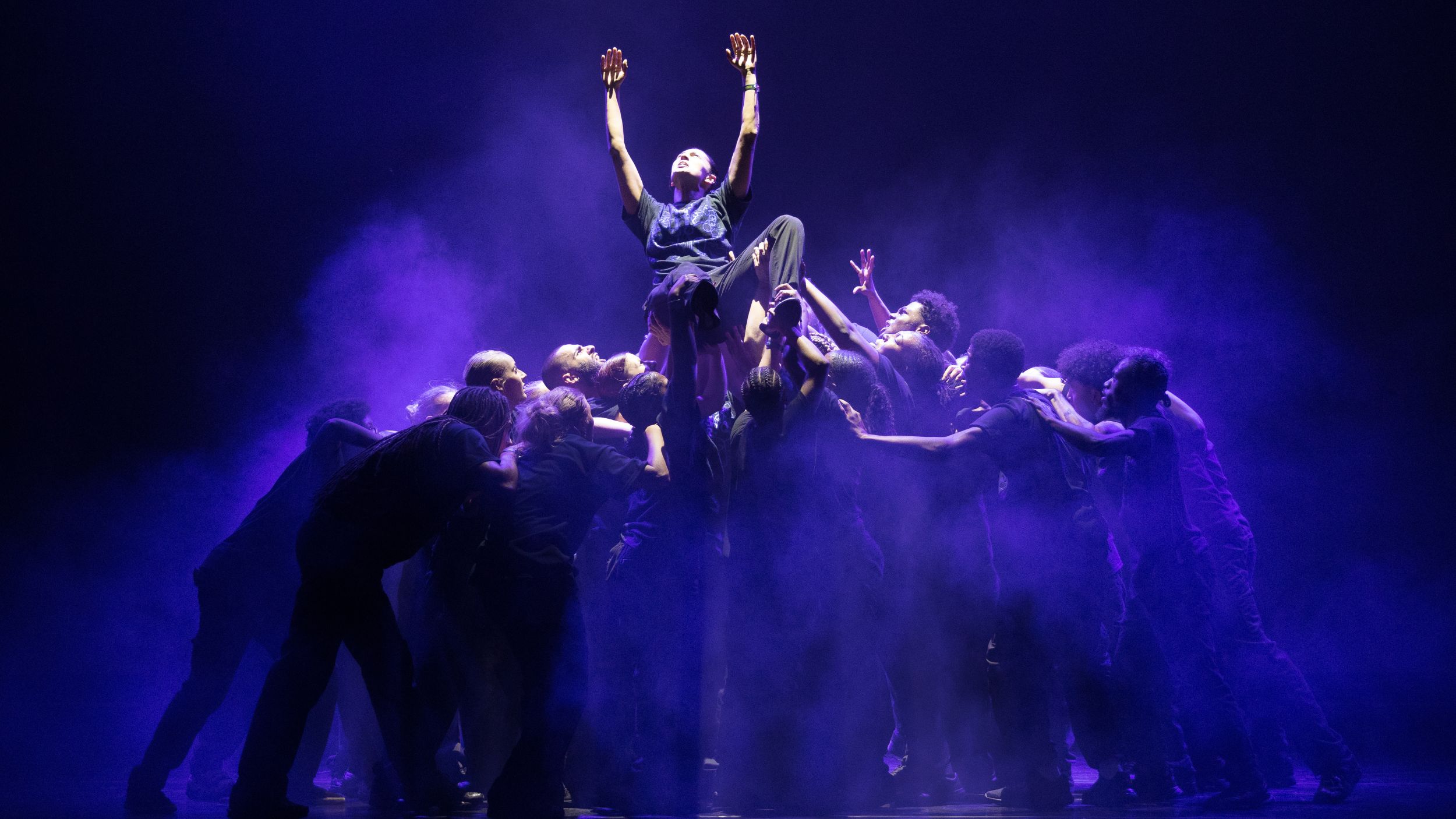 a group of dancers on stage lit in a blue light, one dancer is being lifted up towards the light by the others