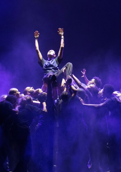 a group of dancers on stage lit in a blue light, one dancer is being lifted up towards the light by the others