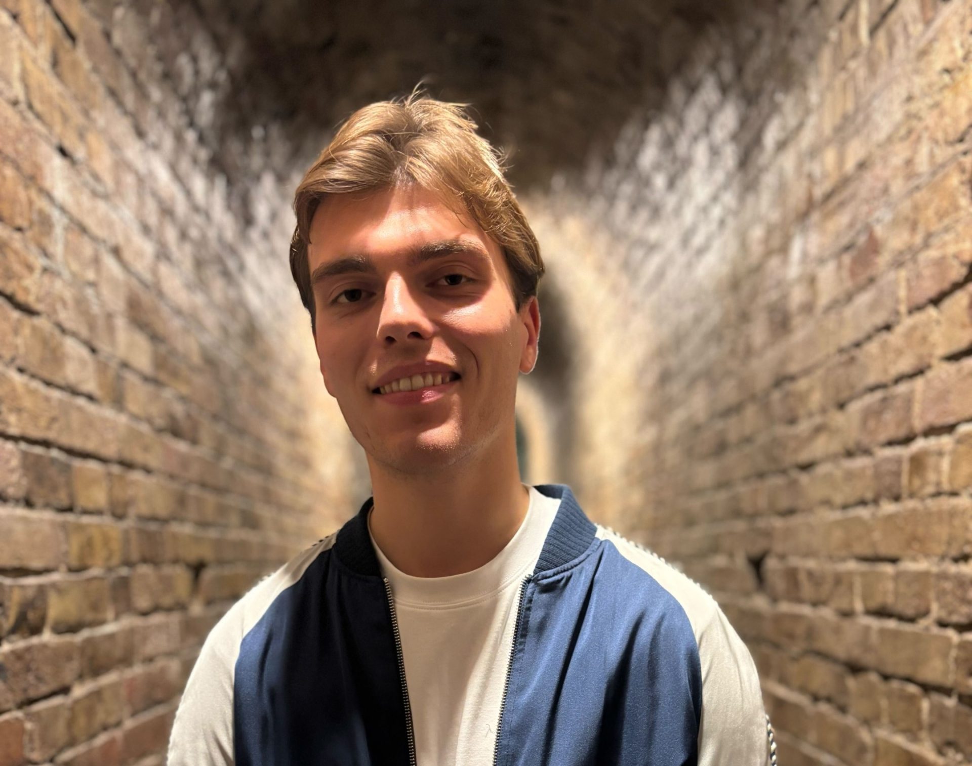 Cameron Goodchild, a smiling young man stands centered in a dimly lit brick archway. He has light brown hair and is wearing a dark blue and white zip-up bomber jacket over a white crew-neck shirt. The brick walls of the tunnel recede into the distance behind him, creating a symmetrical background.