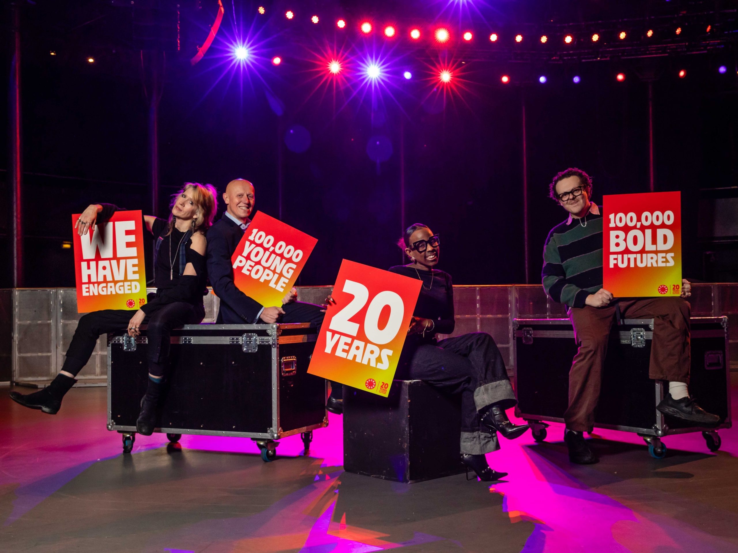 Four people stand in an empty gig venue, holding signs that read 