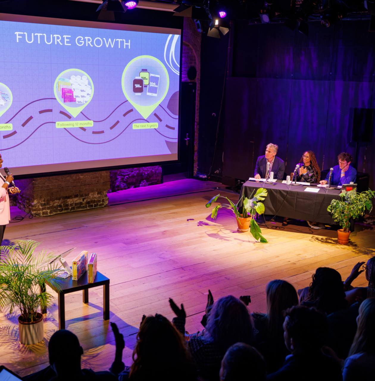 A person standing on a stage presenting in front of a screen with icons, with other people seated at a table and an audience visible in the foreground.