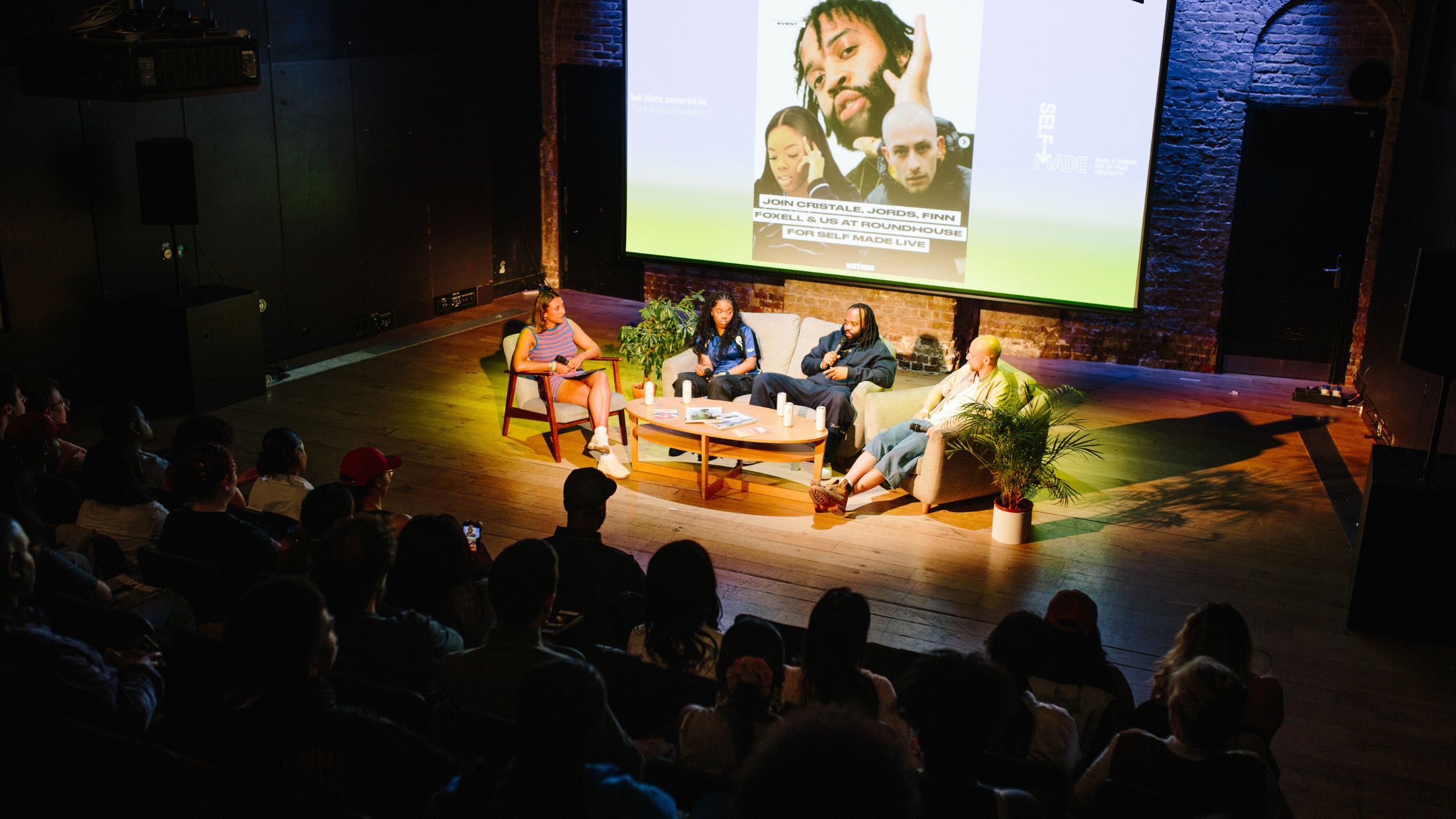 A dimly lit indoor event with an audience, featuring several people on a stage behind a table, and a large screen displaying a person's image above them