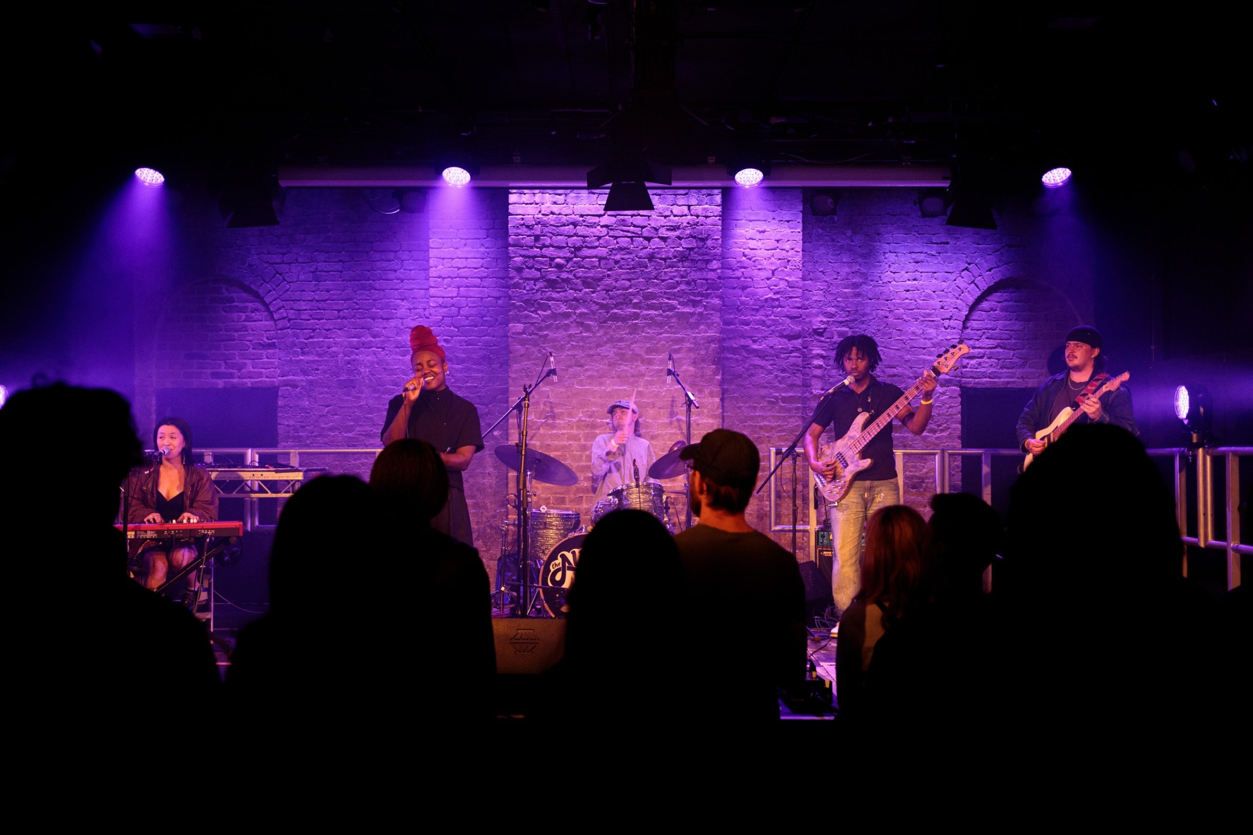 A band performs on a stage in front of a brick background lit by purple lights