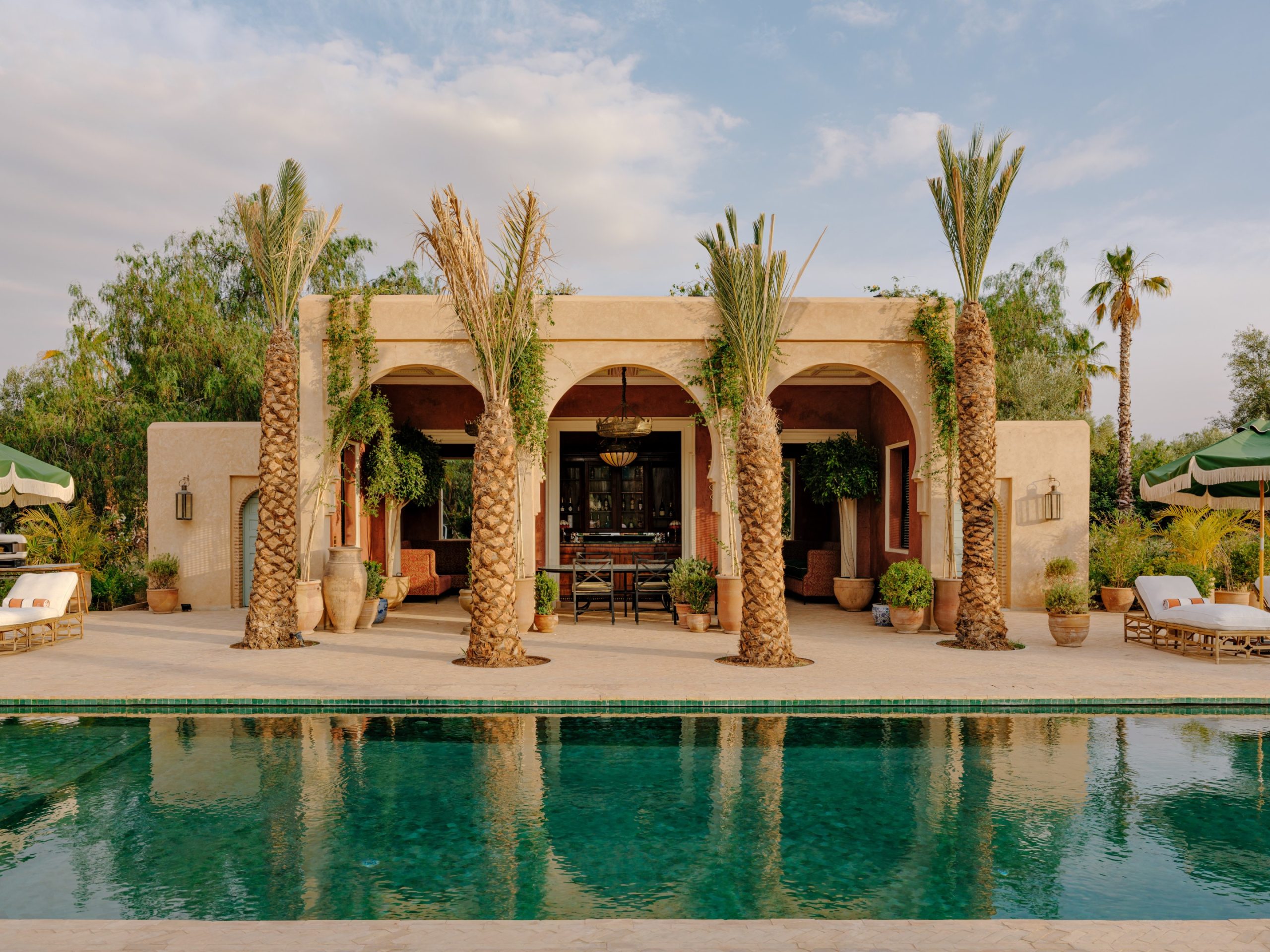 An image of a Moroccan building with three central arches, palm trees and a pool in front.